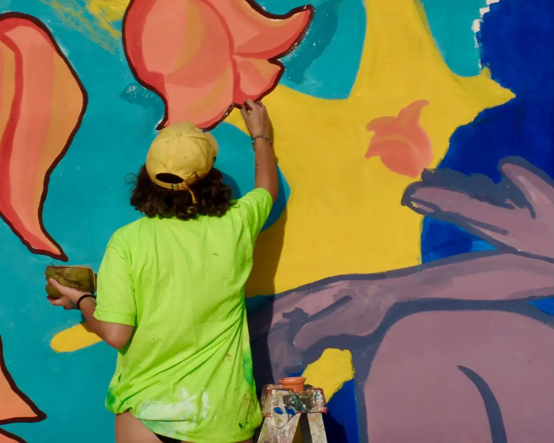 Analía pintando una mano en el mural con gorra amarilla