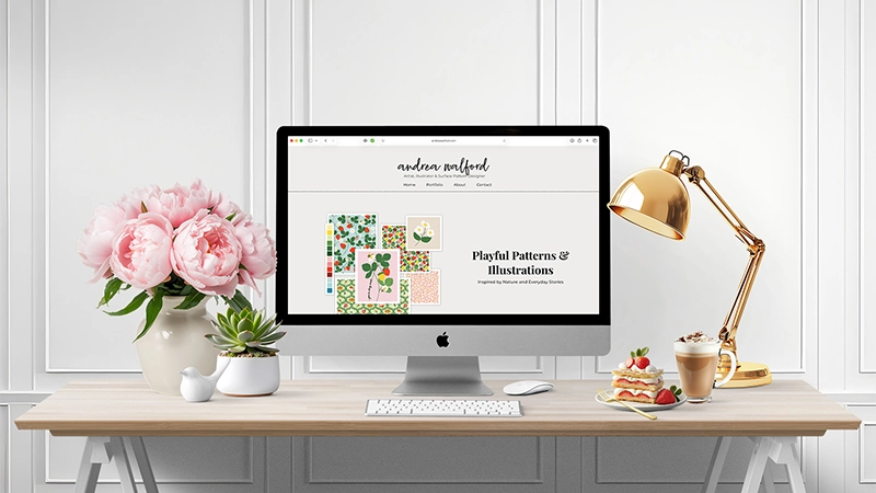 Computer displaying Andrea Walford’s surface pattern design portfolio on a desk beside flowers and a lamp.