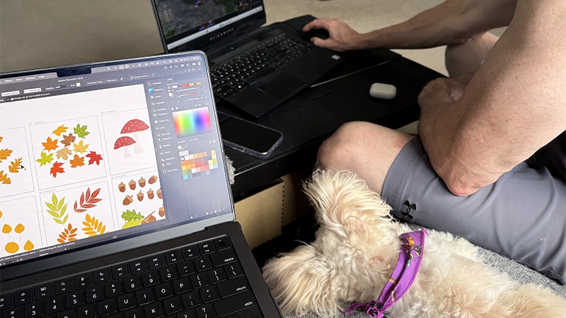 Andrea and her husband relaxing together on a couch while she works on a pattern collection and their dog rests between them.