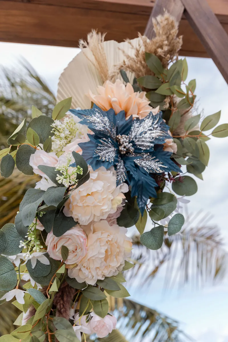 Arreglo floral en tonos melocotón y azul sobre la pérgola con palmas al fondo