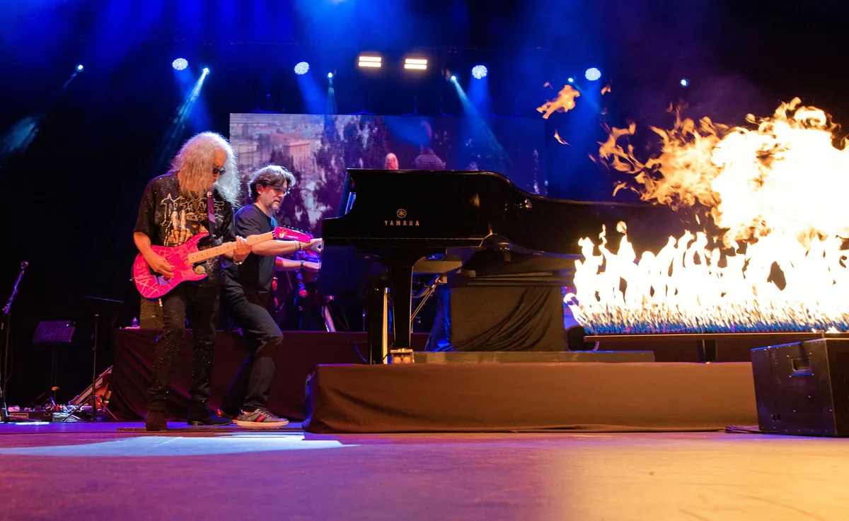 Guitarrista y pianista tocando en un escenario con llamaradas de fuego y luces azules en un concierto