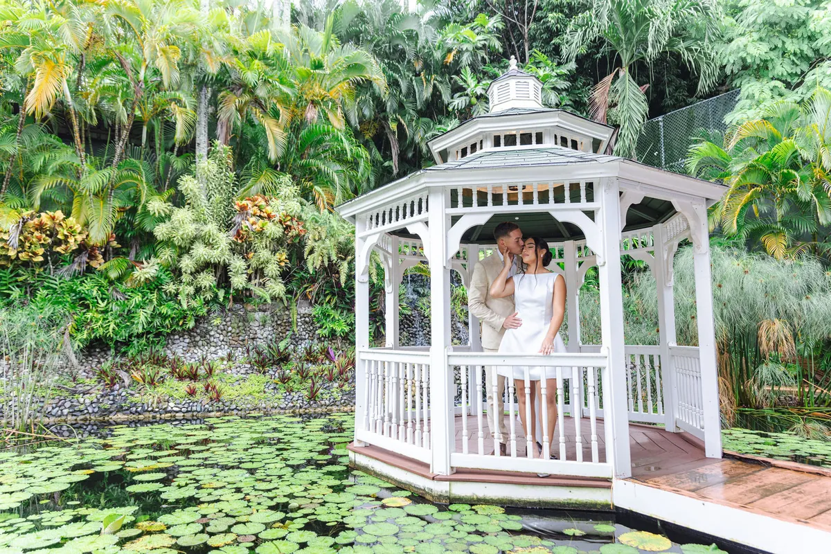 Novios se besan dentro de un gazebo blanco sobre un estanque de lirios rodeado de palmas
