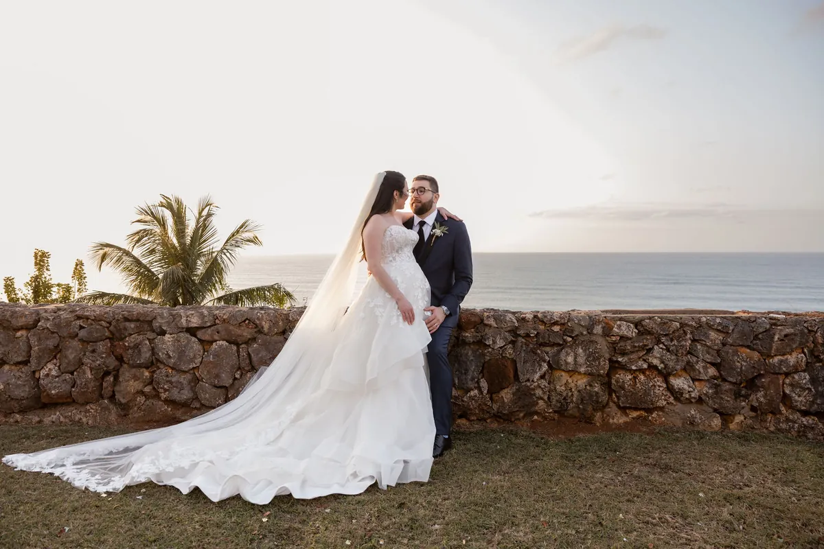 Pareja junto a un muro de piedra frente al mar con cola del vestido extendida y palma