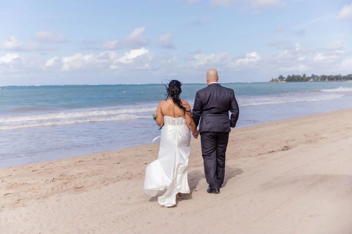 Novios caminan de espaldas por la orilla de la playa bajo un cielo azul con nubes