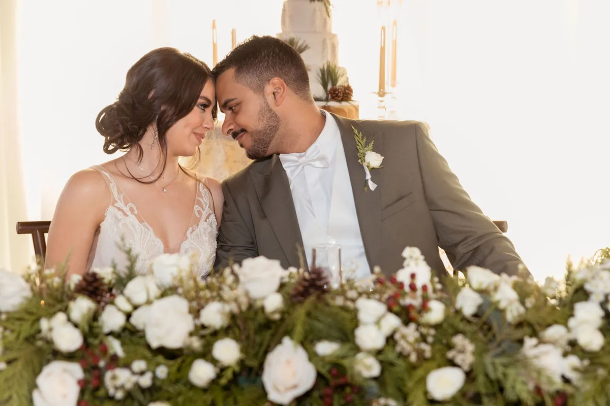 Novios sentados frente con frente tras una mesa de flores blancas y helechos