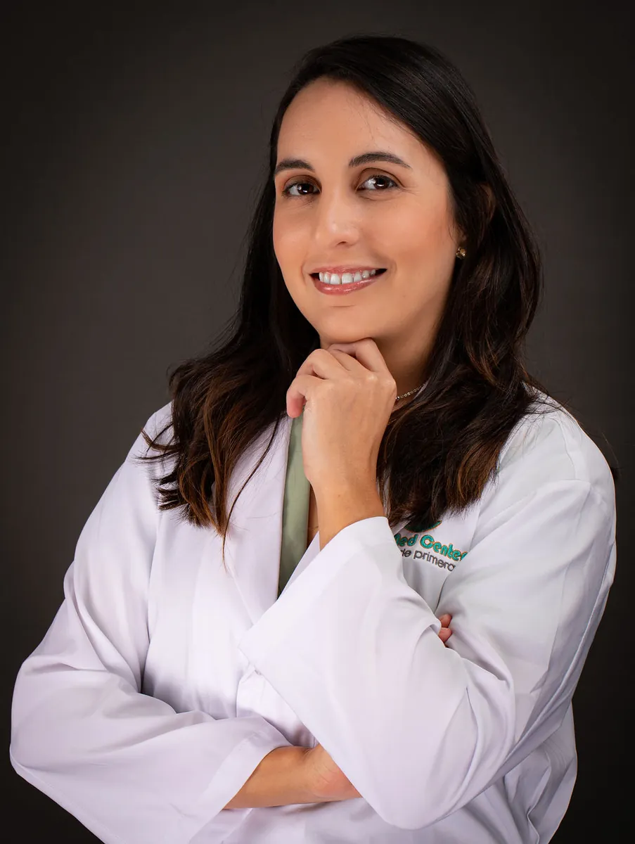 Retrato profesional de una doctora con bata blanca y mano en el mentón sobre fondo gris de estudio