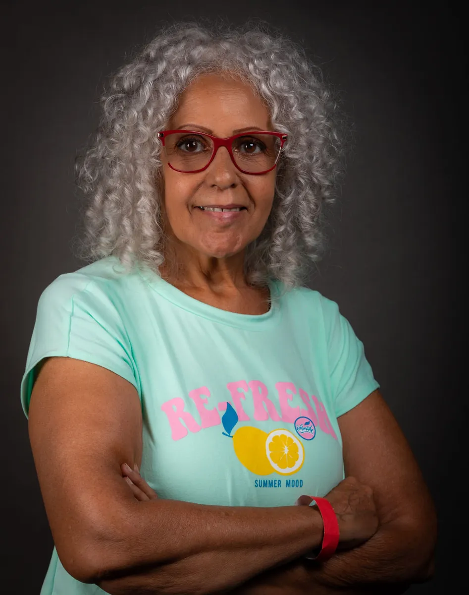 Retrato de estudio de una mujer mayor de cabello gris rizado y espejuelos rojos con brazos cruzados sonriendo