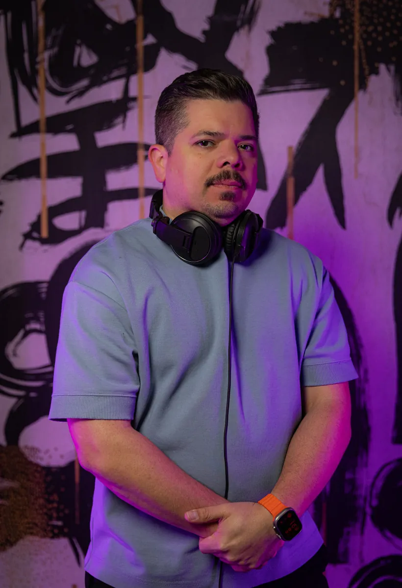 Retrato de un DJ con audífonos al cuello frente a un mural morado de trazos negros