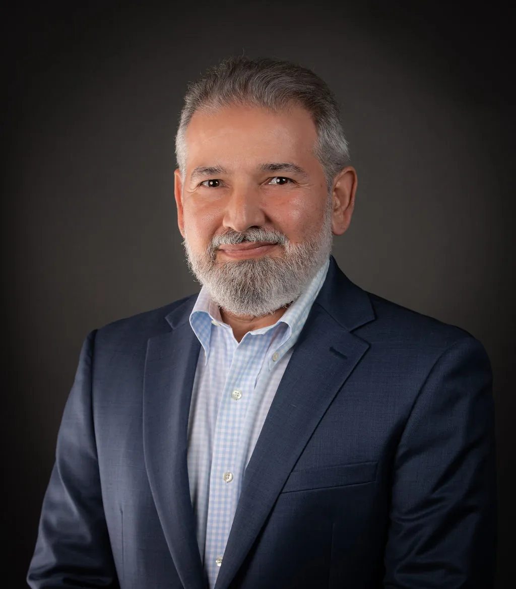 Retrato profesional de un hombre mayor con barba canosa y chaqueta azul marino sobre fondo gris de estudio