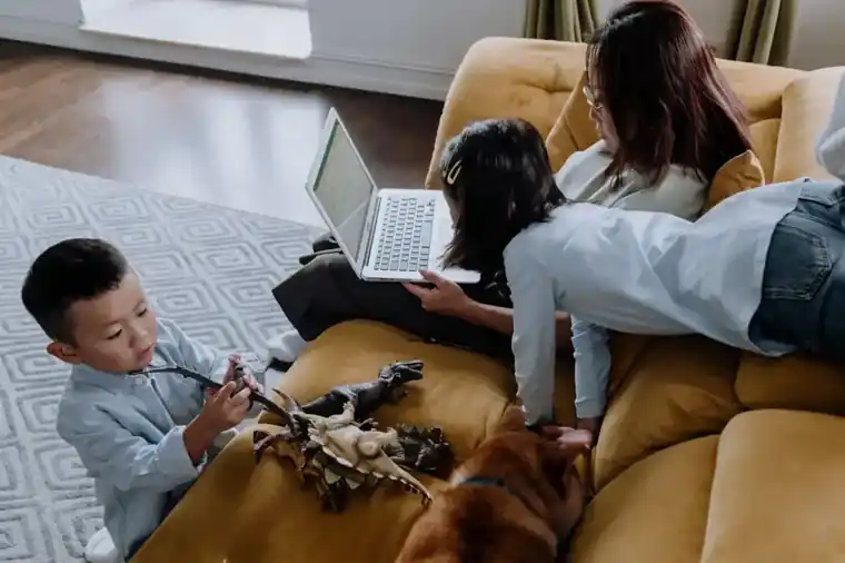 A mom on her laptop on the couch while her kids and dog play around her