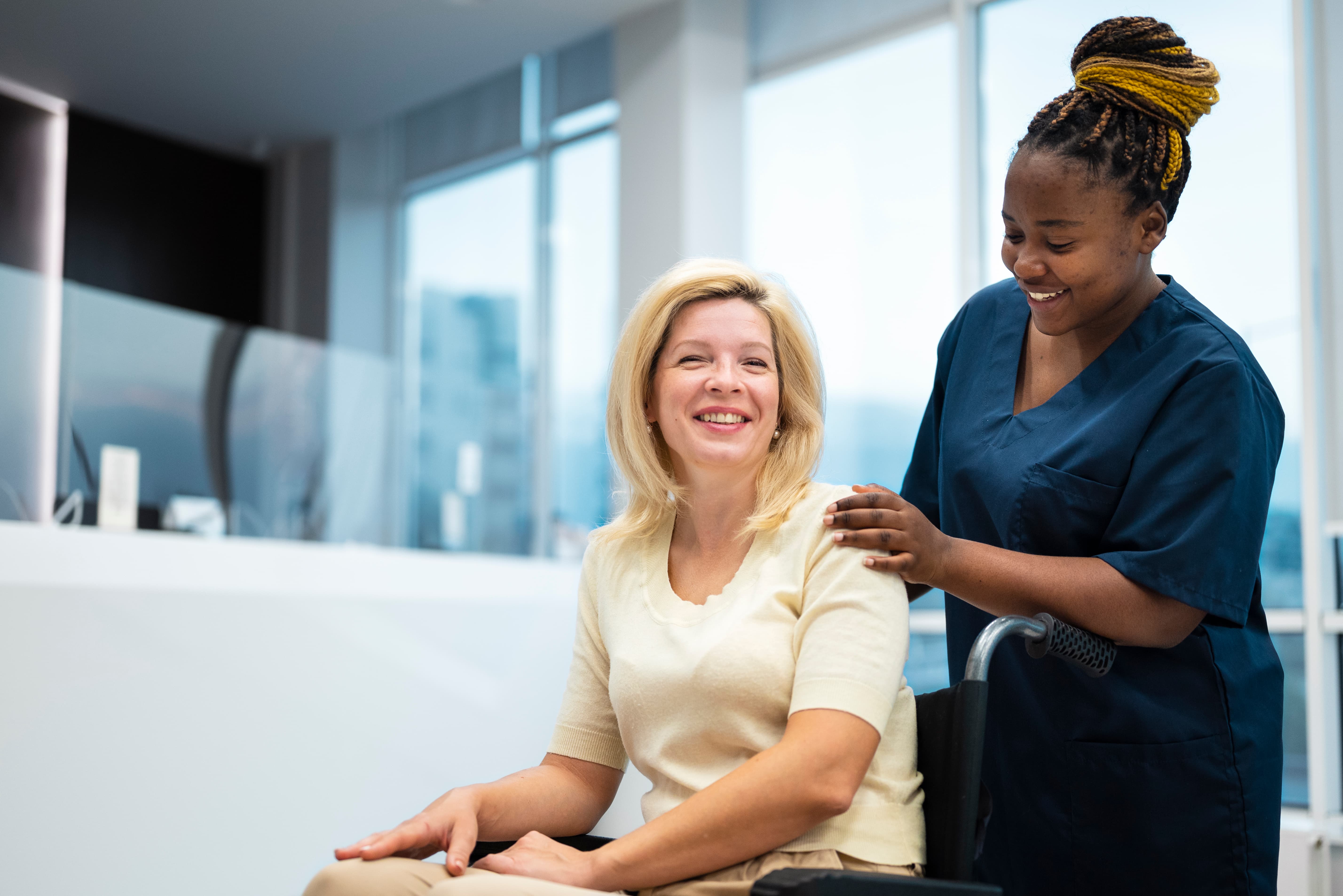 A healthcare professional in dark blue scrubs stands behind a patient seated in a wheelchair, gently resting hands on their shoulders in a bright medical facility with large windows.