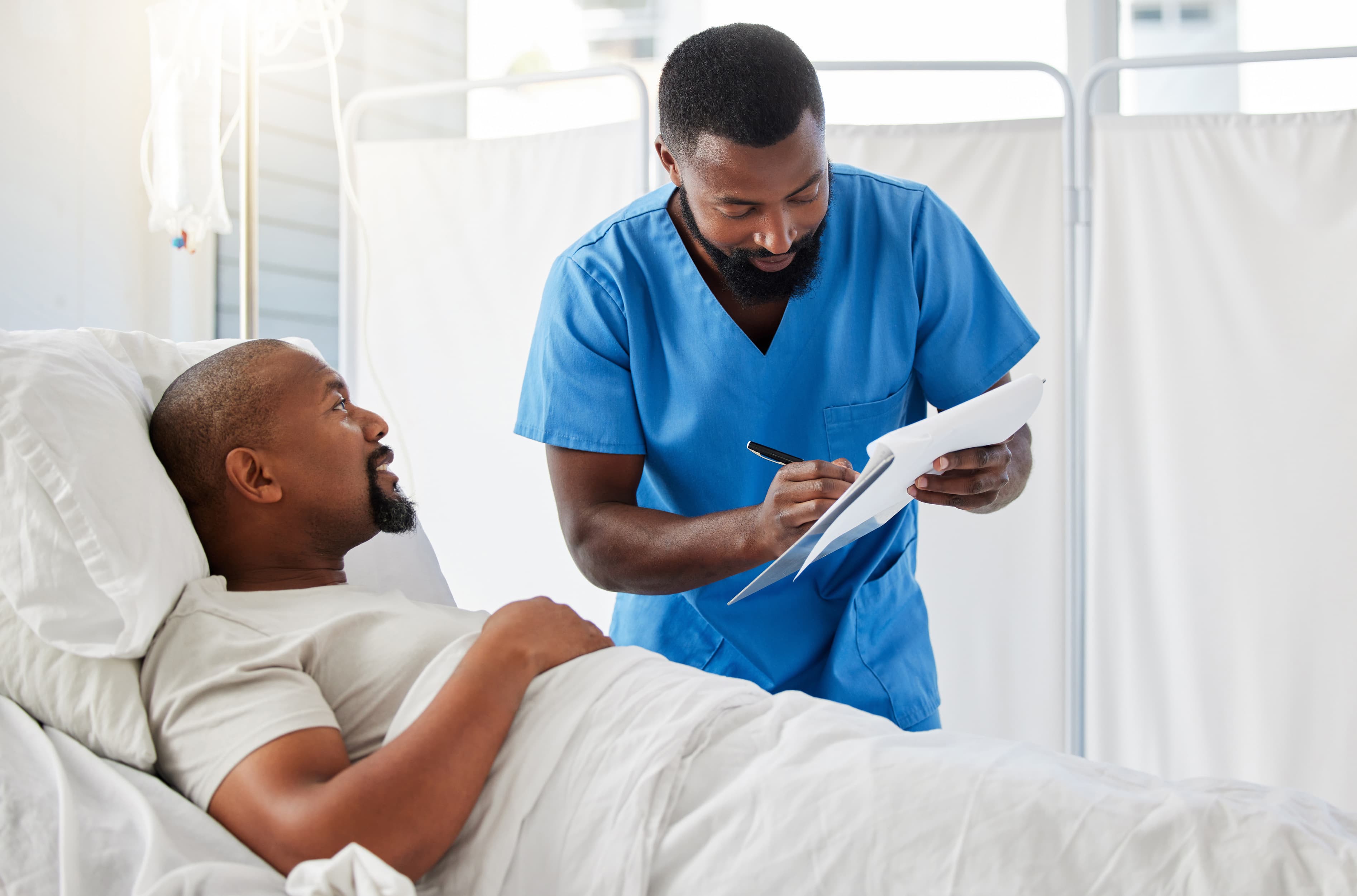 Healthcare professional in blue scrubs writing on a clipboard beside a patient lying in a hospital bed with white sheets in a bright clinical setting. Healthcare professional in blue scrubs writing on a clipboard beside a patient lying in a hospital bed with white sheets in a bright clinical setting.
