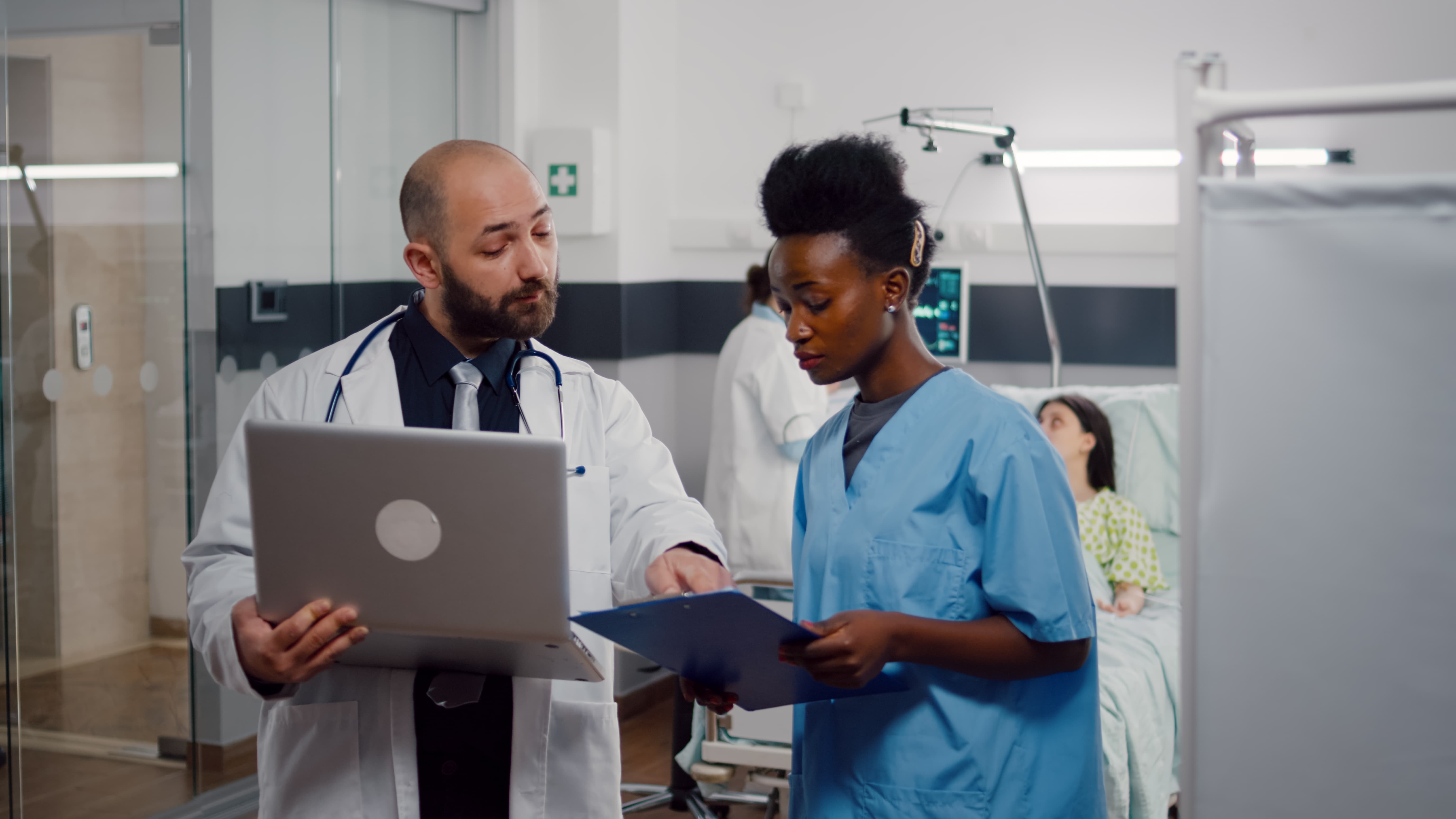 Two healthcare professionals, one in a white coat holding a laptop and another in blue scrubs with a clipboard, discussing patient information beside a hospital bed in a bright modern clinical environment Two healthcare professionals, one in a white coat holding a laptop and another in blue scrubs with a clipboard, discussing patient information beside a hospital bed in a bright modern clinical environment