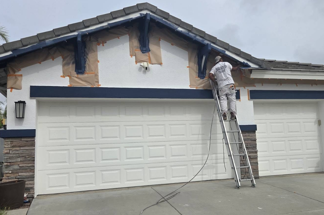 An Upper Room painter on a ladder cutting in navy fascia above a masked-off garage.