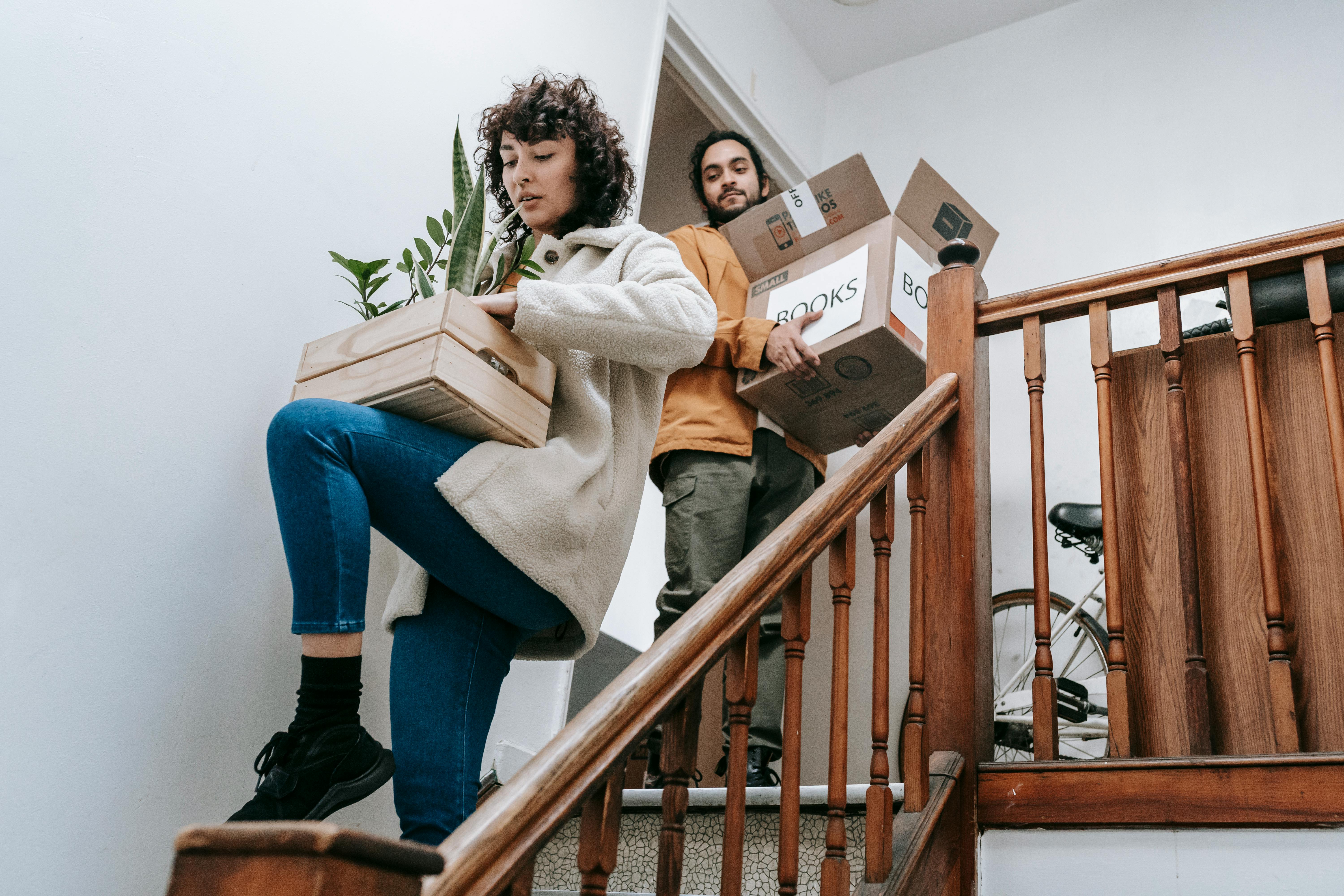 Two individuals appear to be in the process of moving. One person, who seems to be a woman with curly hair, is walking up a wooden staircase carrying two cardboard boxes. She is wearing blue jeans, black socks, and black shoes, along with a light-colored coat. Behind her, on a lower step, a person who appears to be a man with a beard is also carrying a cardboard box. He is wearing a mustard-colored sweater and grey pants. A wooden railing runs along the side of the staircase. In the background, partially visible through a doorway, is a bicycle. A potted plant is also visible on the stairs below the woman. The setting suggests they are moving into or out of a multi-story home.
