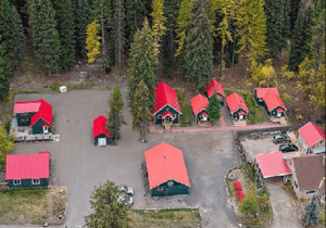   An aerial view shows a cluster of small, dark-colored buildings with bright red roofs nestled among tall evergreen and some yellowing deciduous trees. The buildings appear to be cabins or small houses, scattered across a mostly gravel or dirt ground. Some of the buildings have small porches or entryways. A few parked vehicles are visible near some of the structures. The density of the trees suggests a wooded or forested area. The overall scene conveys a sense of a remote or secluded lodging area, possibly a resort or a small community within a natural environment.