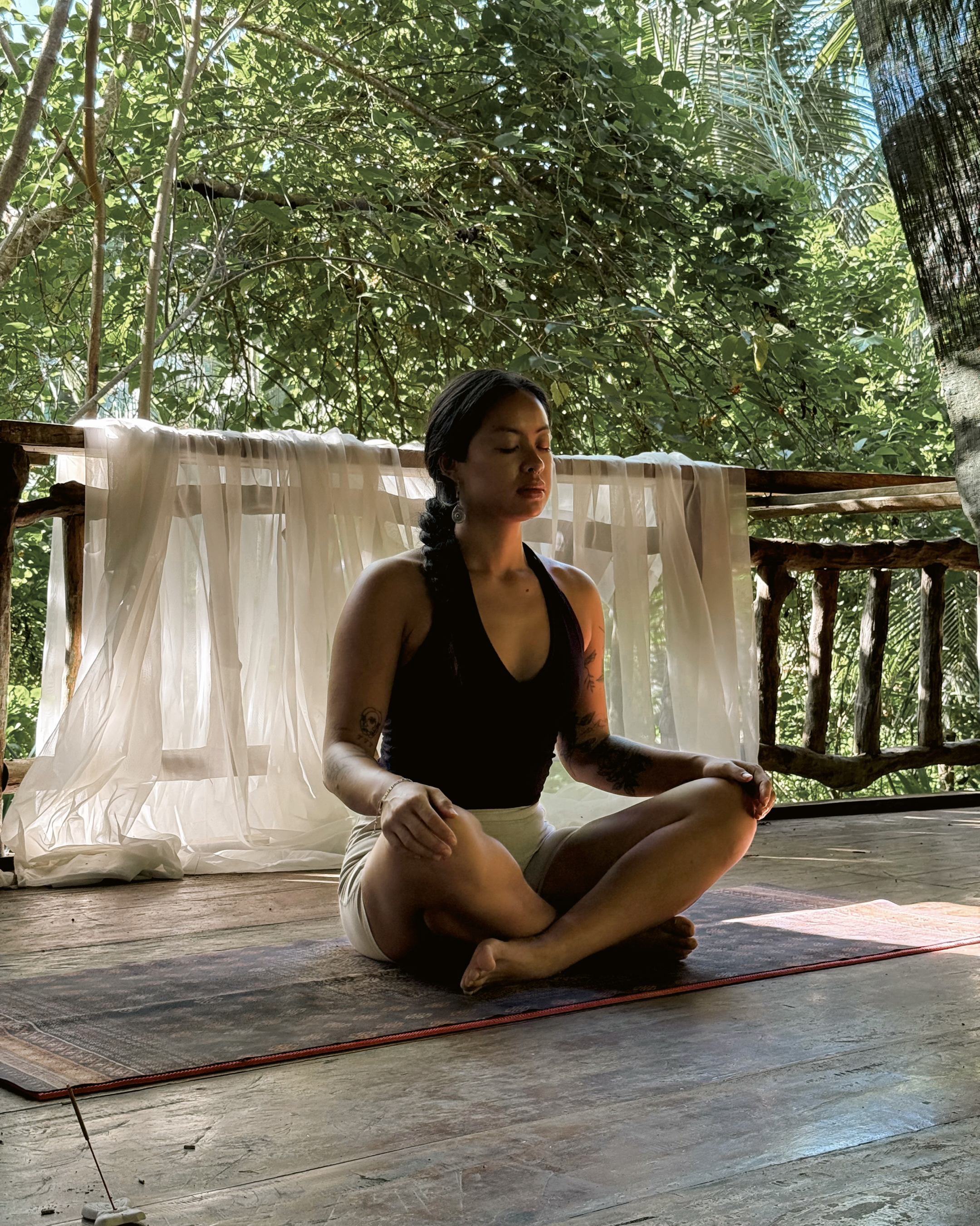 Guest in seated meditation on the farm deck, sheer curtains behind her, jungle canopy in the background