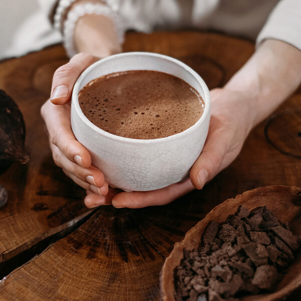 Hands cradling a white cup of warm cacao on a wooden slab, cacao nibs and a pod beside it