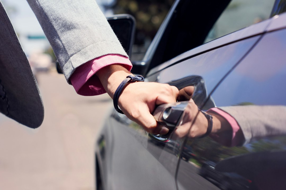 Close-up of a hand in a grey suit sleeve opening the door of a dark sedan on a sunny street.