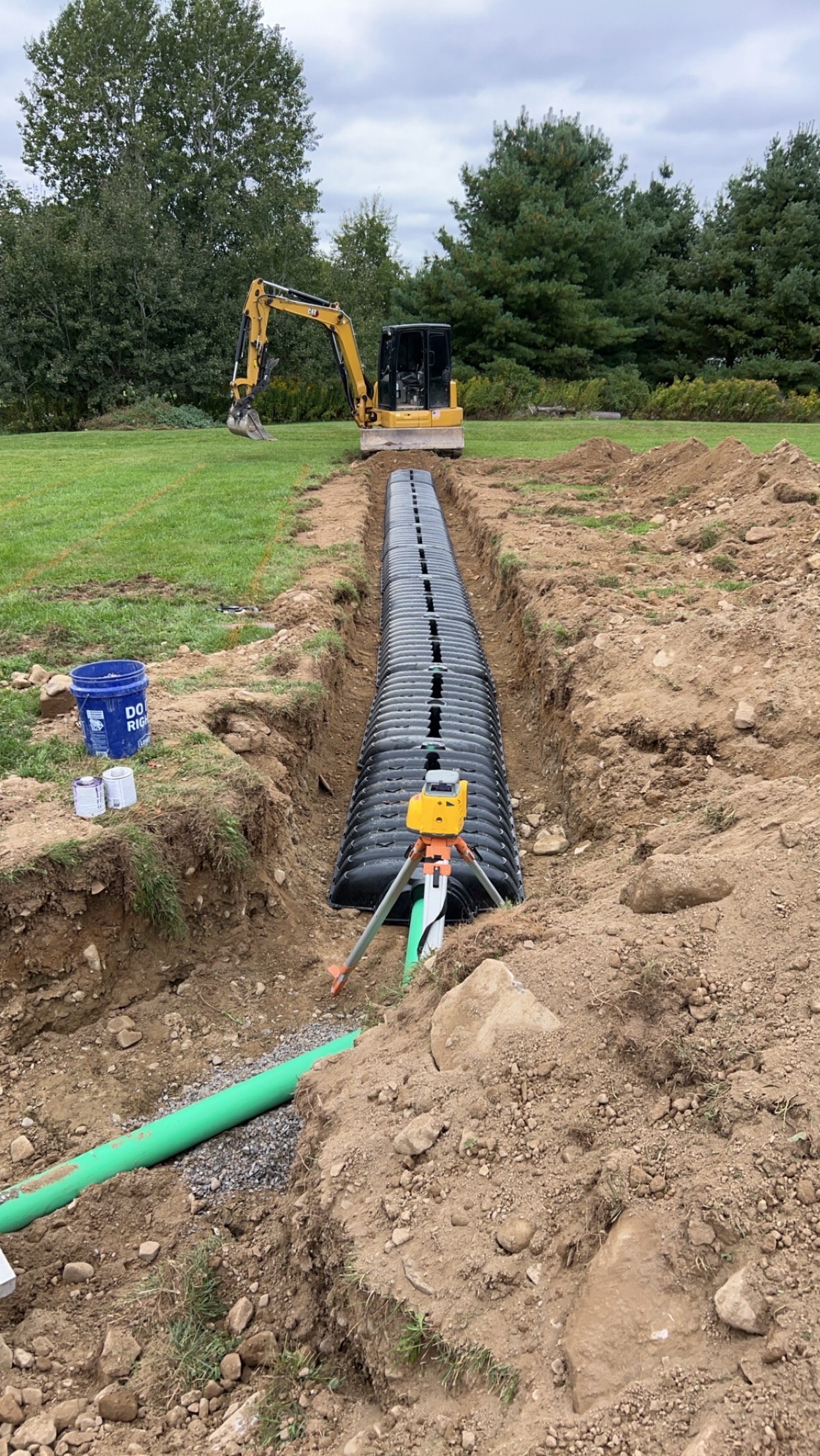 Excavator digging a septic system trench on a residential lot in the Lansing, Michigan area, with clay soil visible along the trench walls. Drain field pipe sections are staged nearby on the surface, ready for installation.