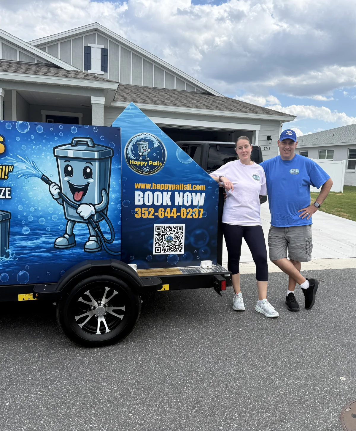 A man and woman standing by a "Happy Pails" trash can cleaning service trailer in front of a house.
