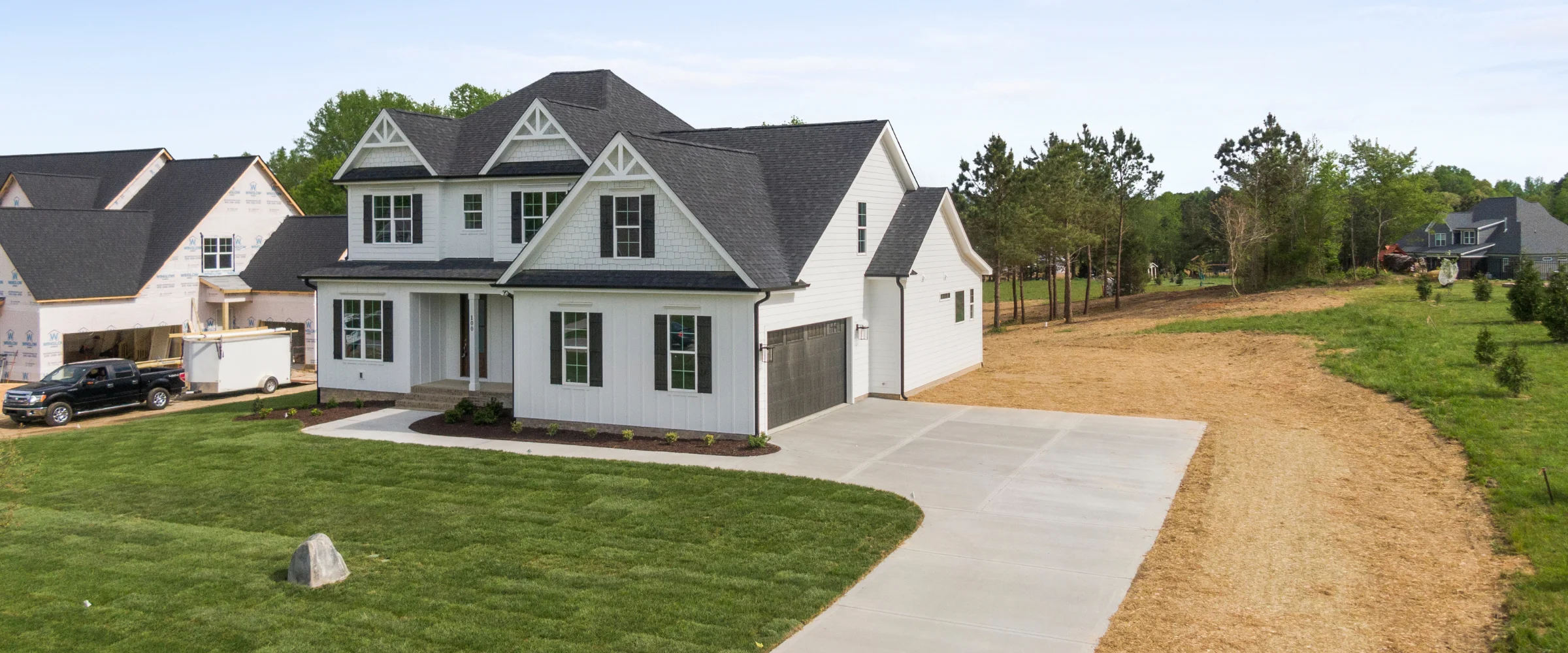 A newly built white house with black roof and shutters, flanked by a construction site and barren land.