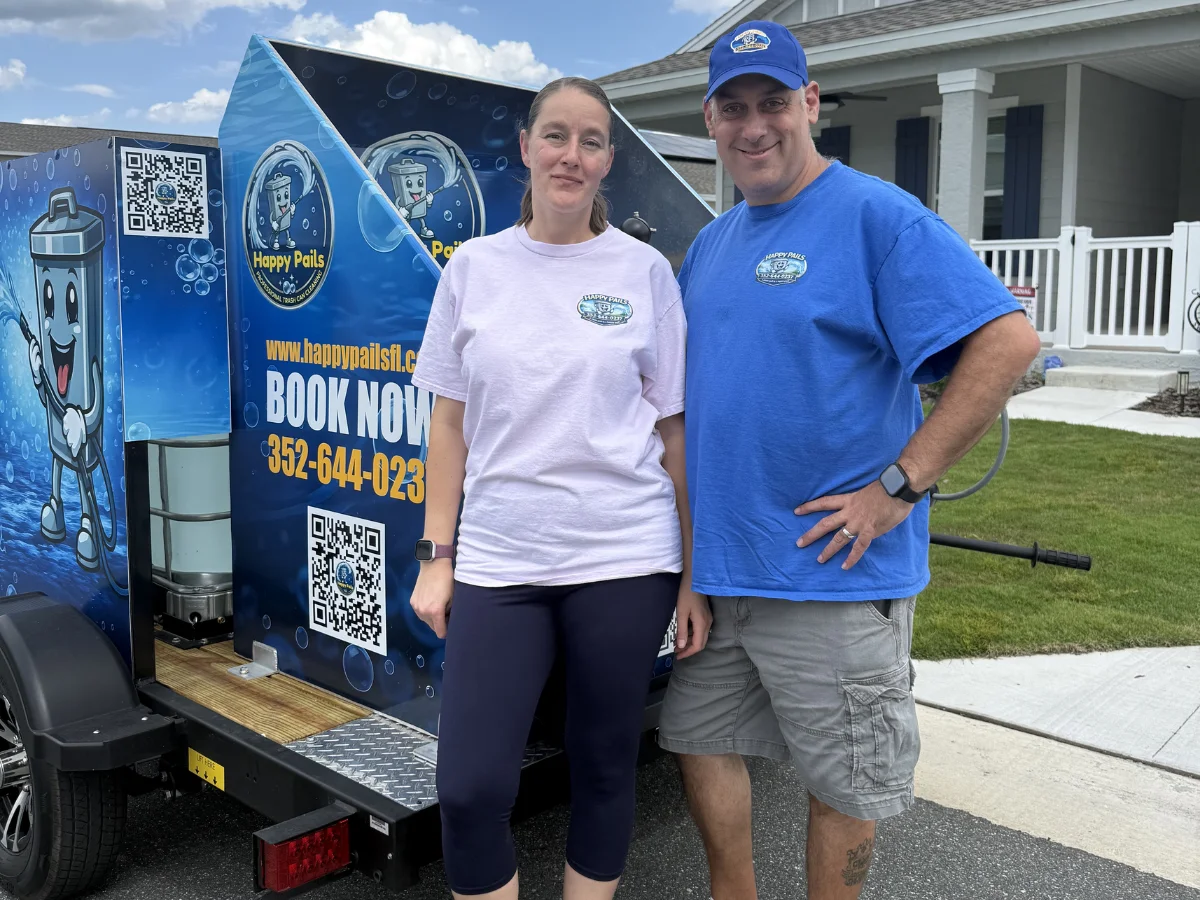 A man and a woman, owners of Happy Pails, stand proudly beside their custom trash can cleaning trailer.