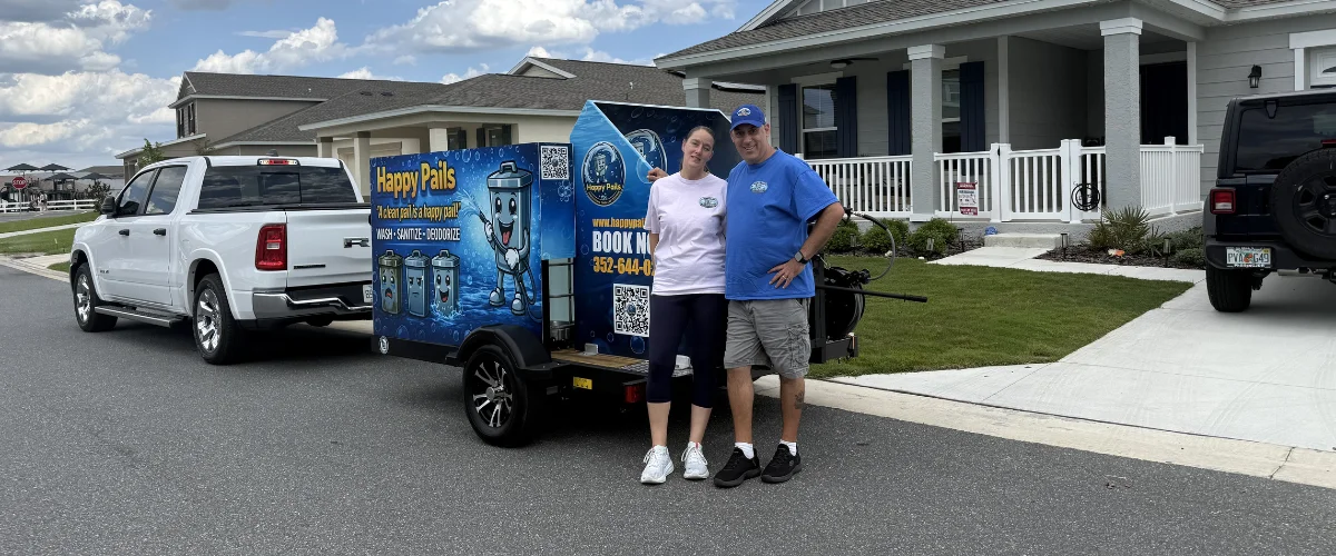 A man and woman standing next to a Happy Pails branded trailer, with a white pickup truck, on a residential street.