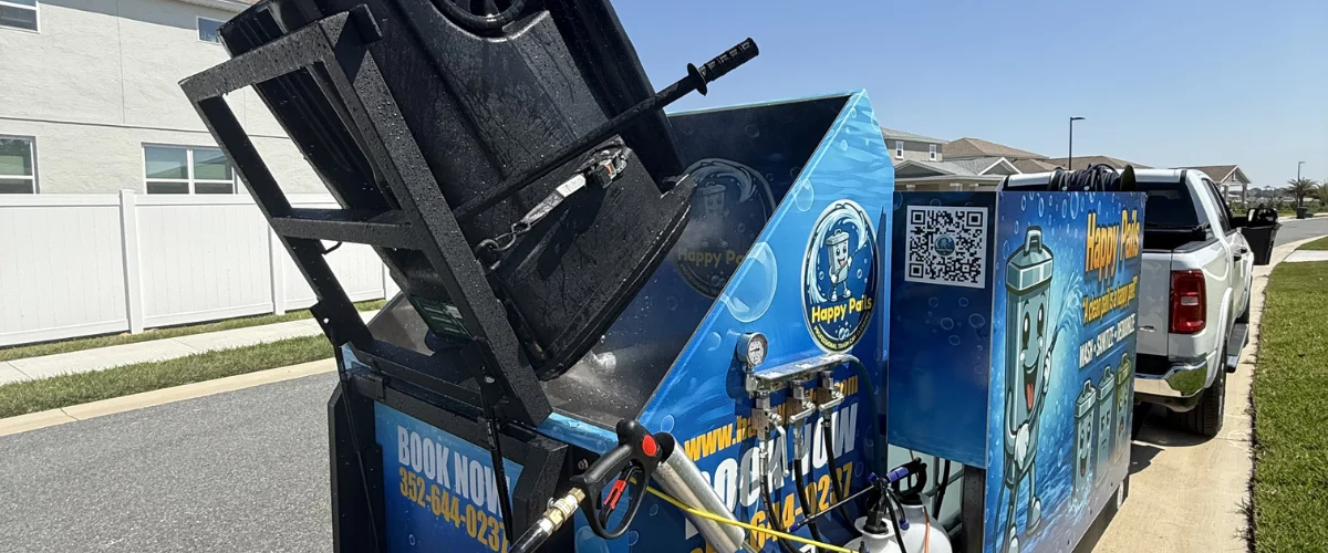 A mobile garbage bin cleaning system with a bin tilted inside, parked next to a white pickup truck on a sunny street.