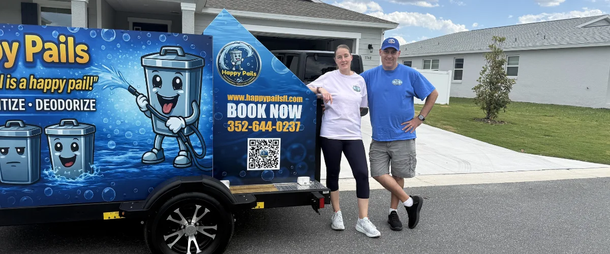 A man and woman smiling next to a branded trailer with the Happy Pails logo and contact information on a residential street.