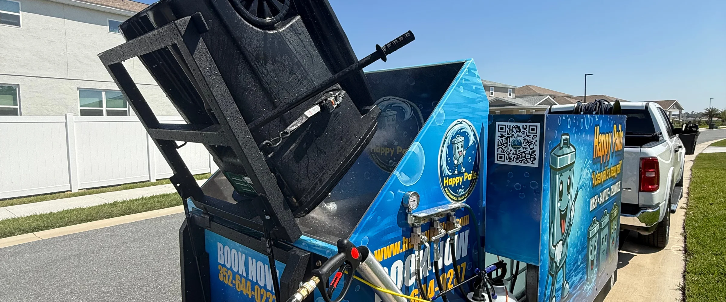 A black trash can being cleaned by a blue Happy Pails professional cleaning unit attached to a white truck.