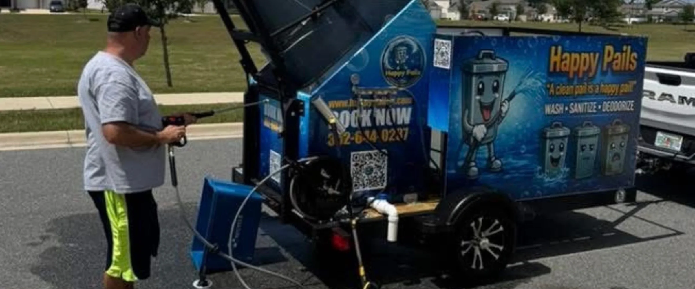 A man holds a pressure washer next to a blue 'Happy Pails' trash can cleaning trailer with cartoon trash cans. A man holds a pressure washer next to a blue 'Happy Pails' trash can cleaning trailer with cartoon trash cans.