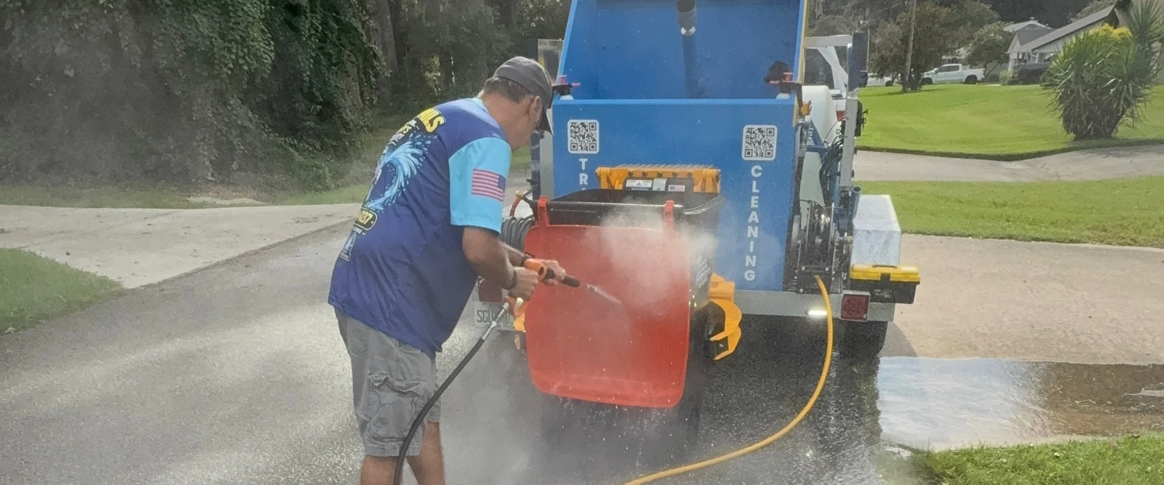 A man in a blue shirt with an American flag patch pressure washing a red garbage bin on a residential street. A man in a blue shirt with an American flag patch pressure washing a red garbage bin on a residential street.