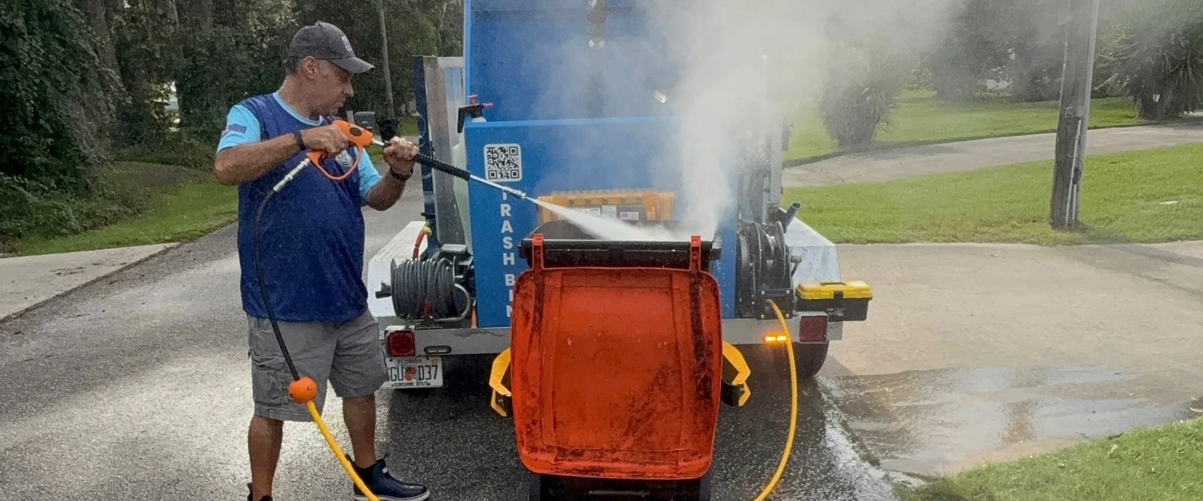 A man in a blue shirt pressure washing an orange trash bin with a mobile cleaning unit on a residential street. A man in a blue shirt pressure washing an orange trash bin with a mobile cleaning unit on a residential street.