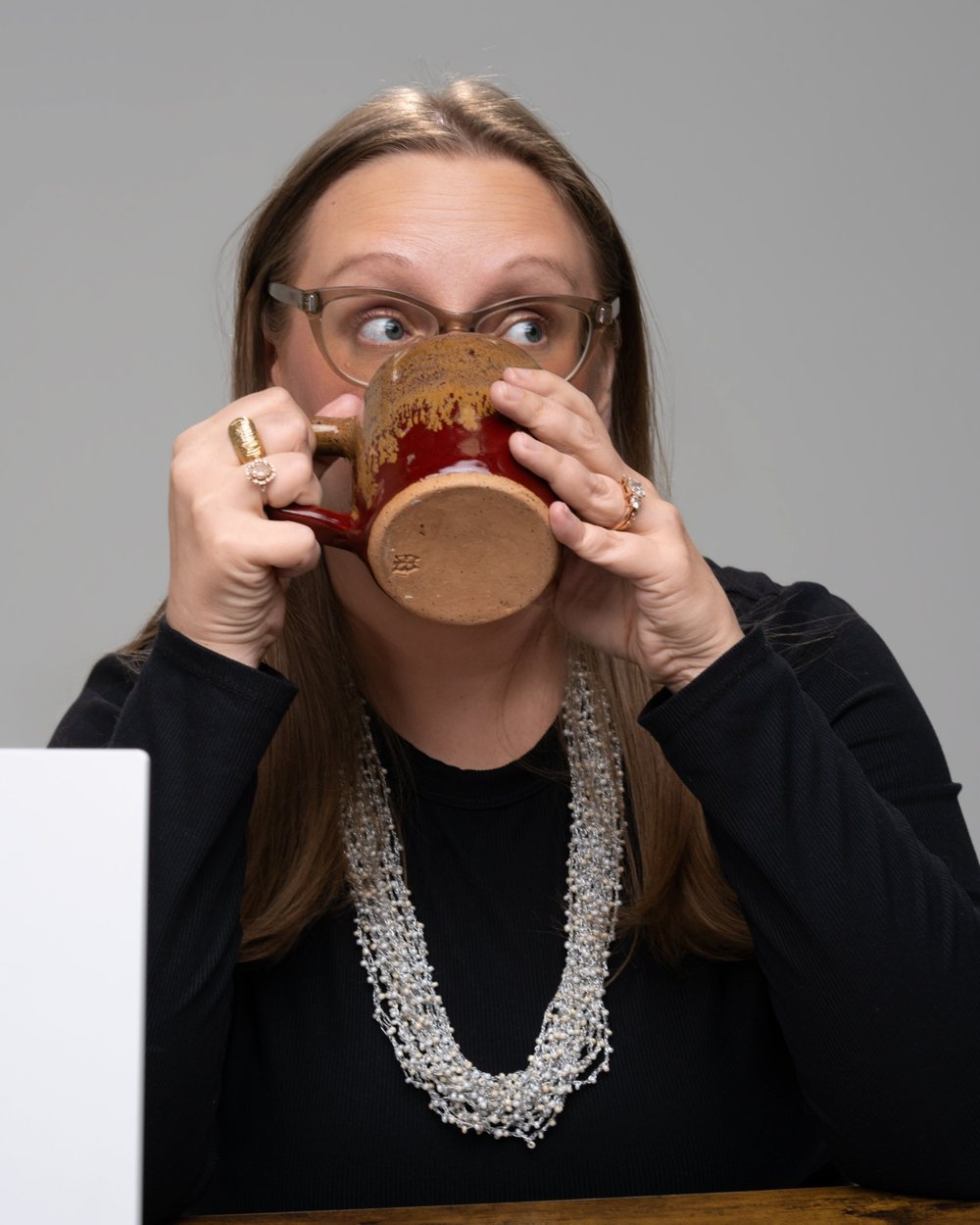Tara taking a big drink from a mug and glancing sideways, laptop beside her