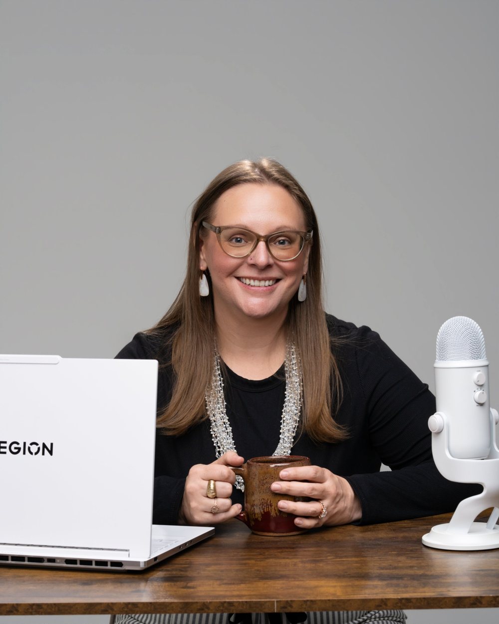 Tara smiling at her desk, holding a mug, with a laptop and microphone beside her