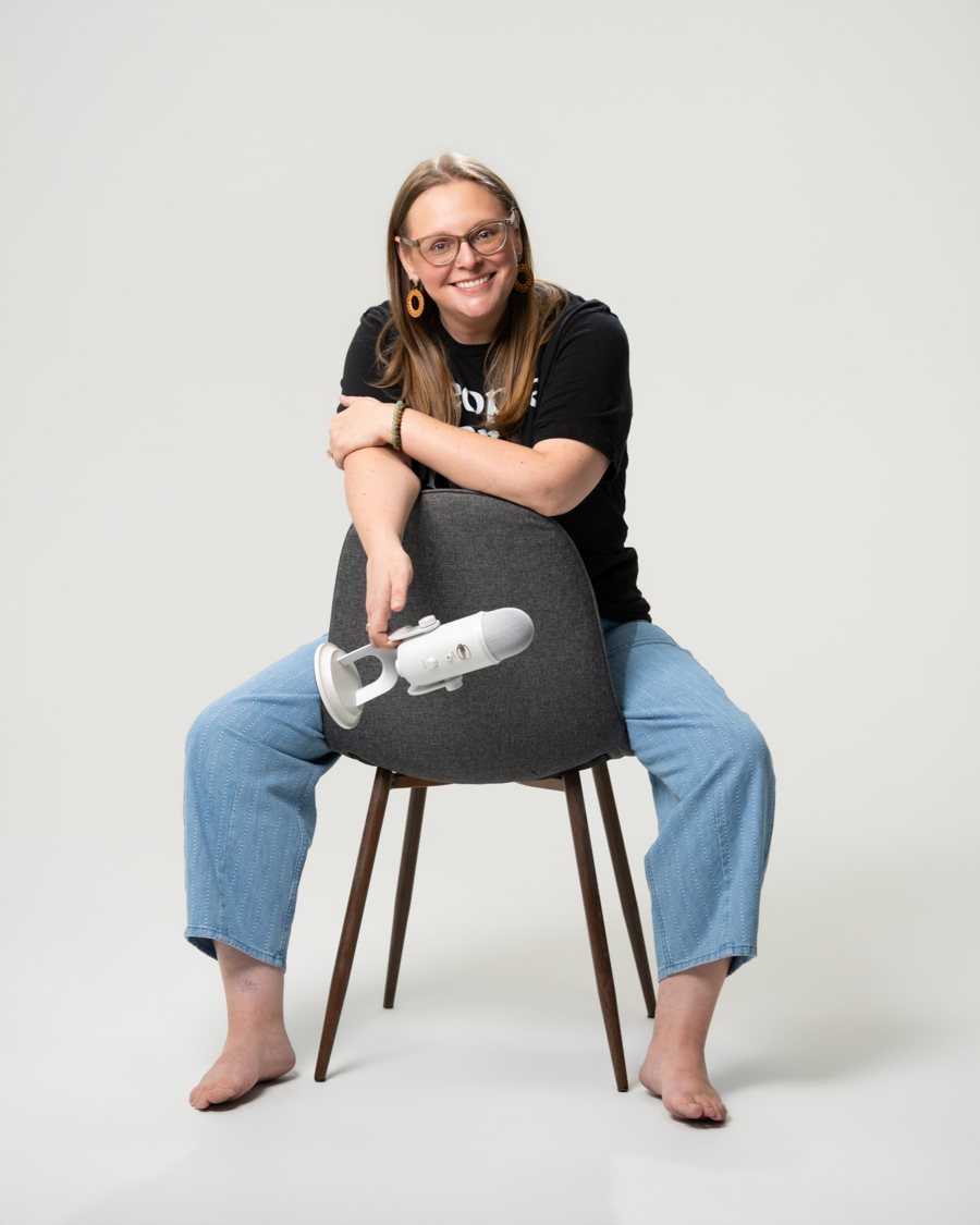 Tara sitting backwards on a chair, smiling, holding a white microphone low beside her