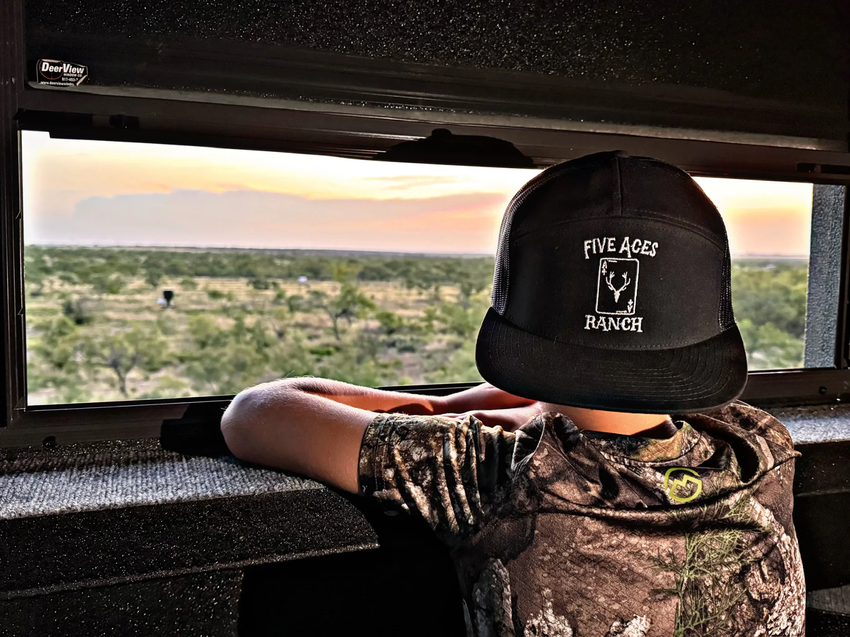 Young hunter in a Five Aces Ranch cap looking out a hunting blind window over Texas brush country at sunset