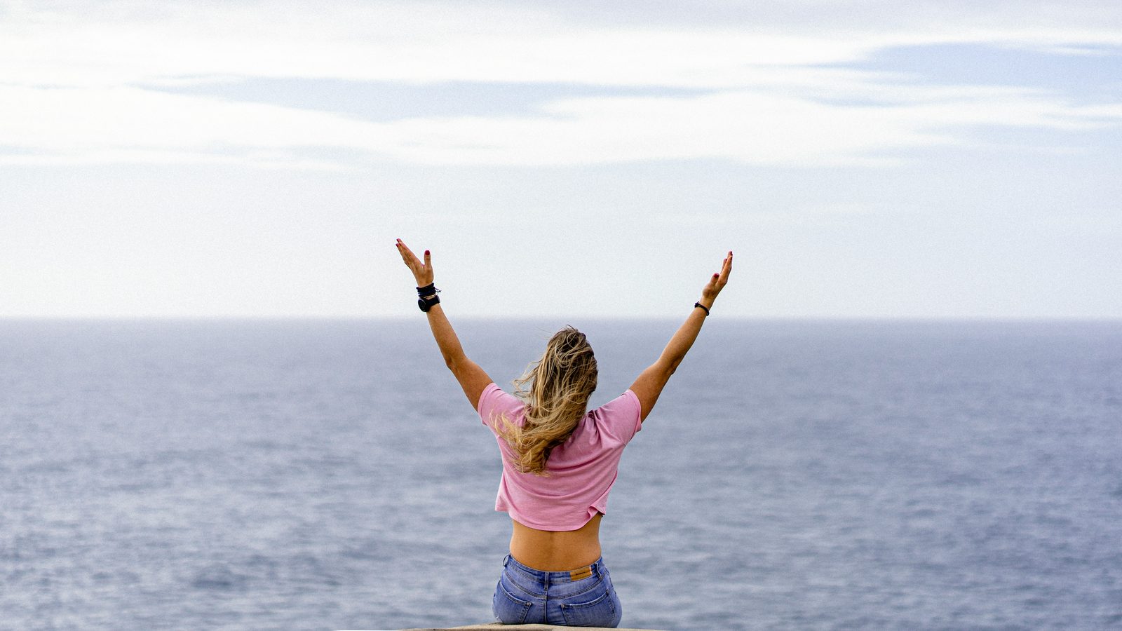 A woman sitting at the water's edge with both arms raised toward a wide open sky