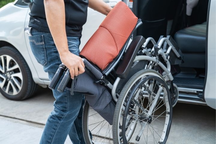 A person folding a portable model next to a car to demonstrate the storage of various wheelchair types. A person folding a portable model next to a car to demonstrate the storage of various wheelchair types.