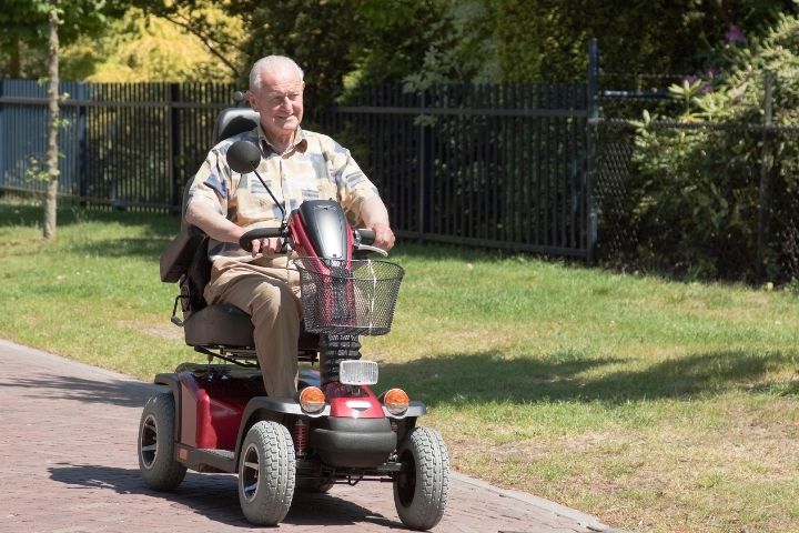 An elderly man driving a red mobility scooter along a scenic paved outdoor path. An elderly man driving a red mobility scooter along a scenic paved outdoor path.