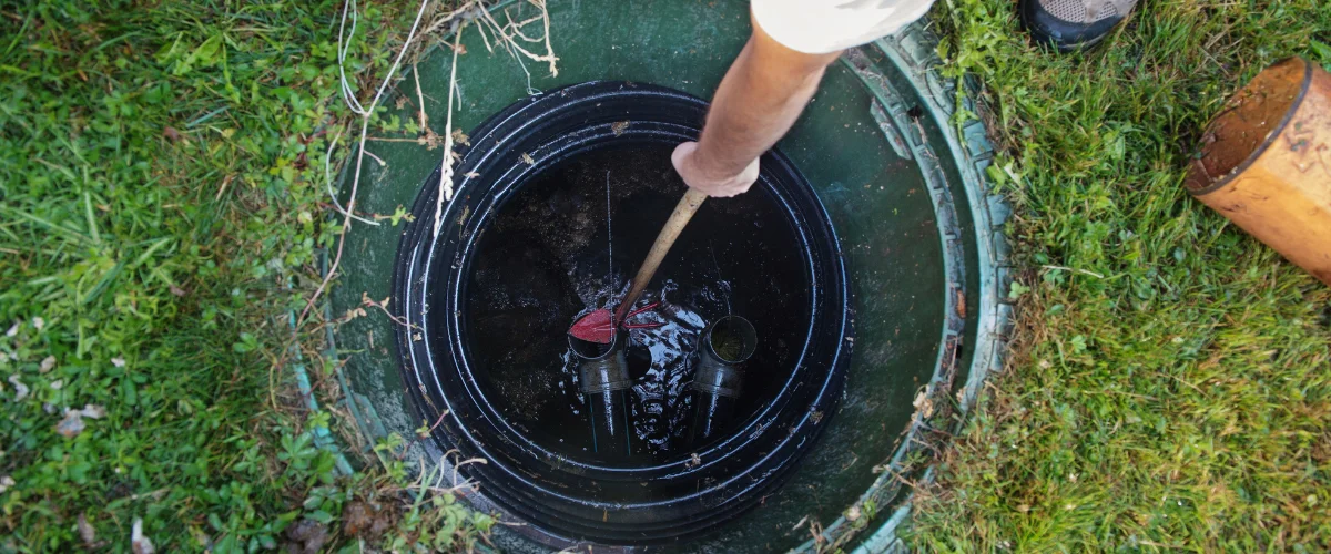 A person uses a tool to inspect the inside of a dark, open septic tank surrounded by green grass.