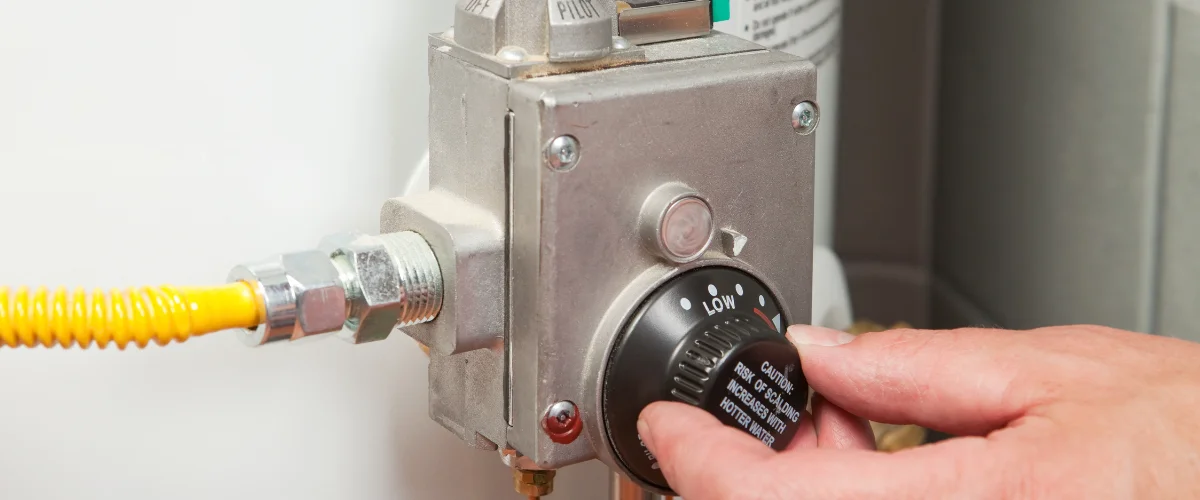 A person's hand adjusts the black temperature dial on a water heater, with a yellow gas line visible.