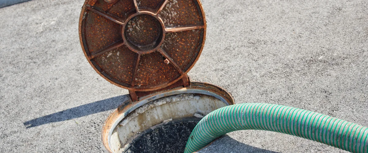 A green corrugated hose extends into an open, rusty manhole cover on a grey asphalt surface, for septic tank maintenance.