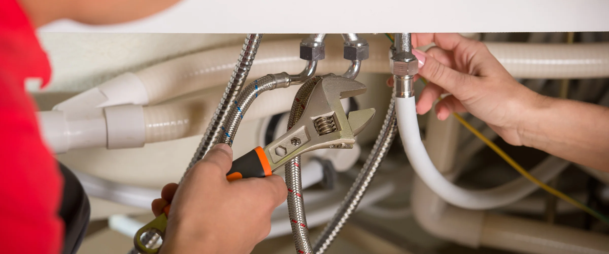 Two people working on plumbing, one using a wrench on a braided hose, the other adjusting a white pipe connection.
