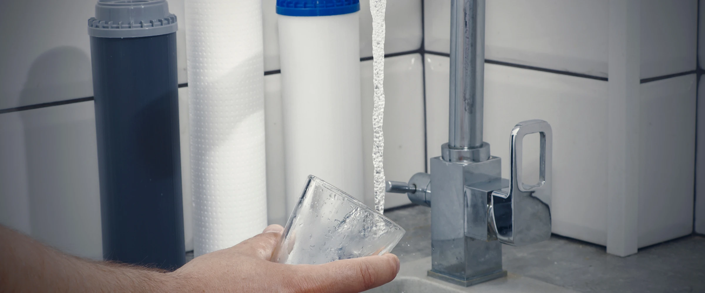 A hand holds a glass under running water from a modern tap, with two household water filters visible in the background. A hand holds a glass under running water from a modern tap, with two household water filters visible in the background.