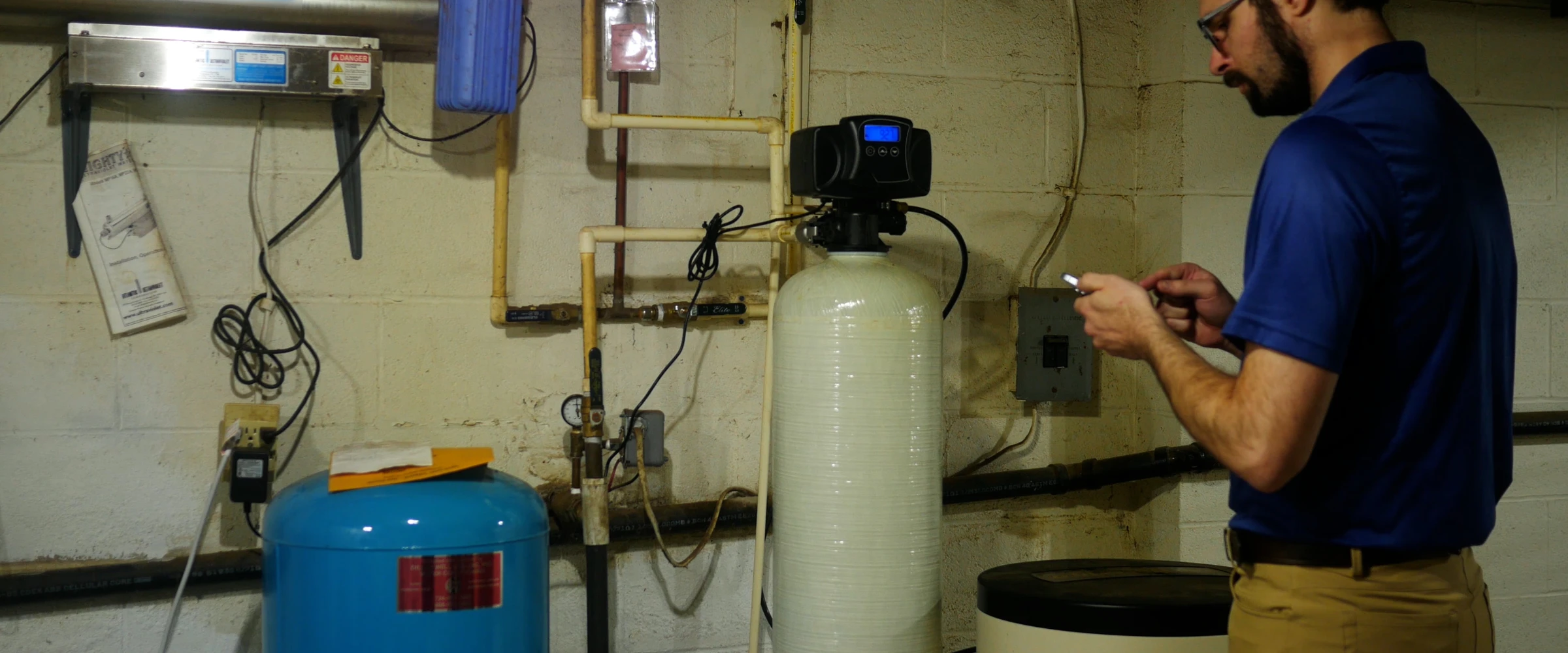 A man in a blue shirt and khakis adjusts settings on a water filtration system in a basement next to pipes and tanks. A man in a blue shirt and khakis adjusts settings on a water filtration system in a basement next to pipes and tanks.