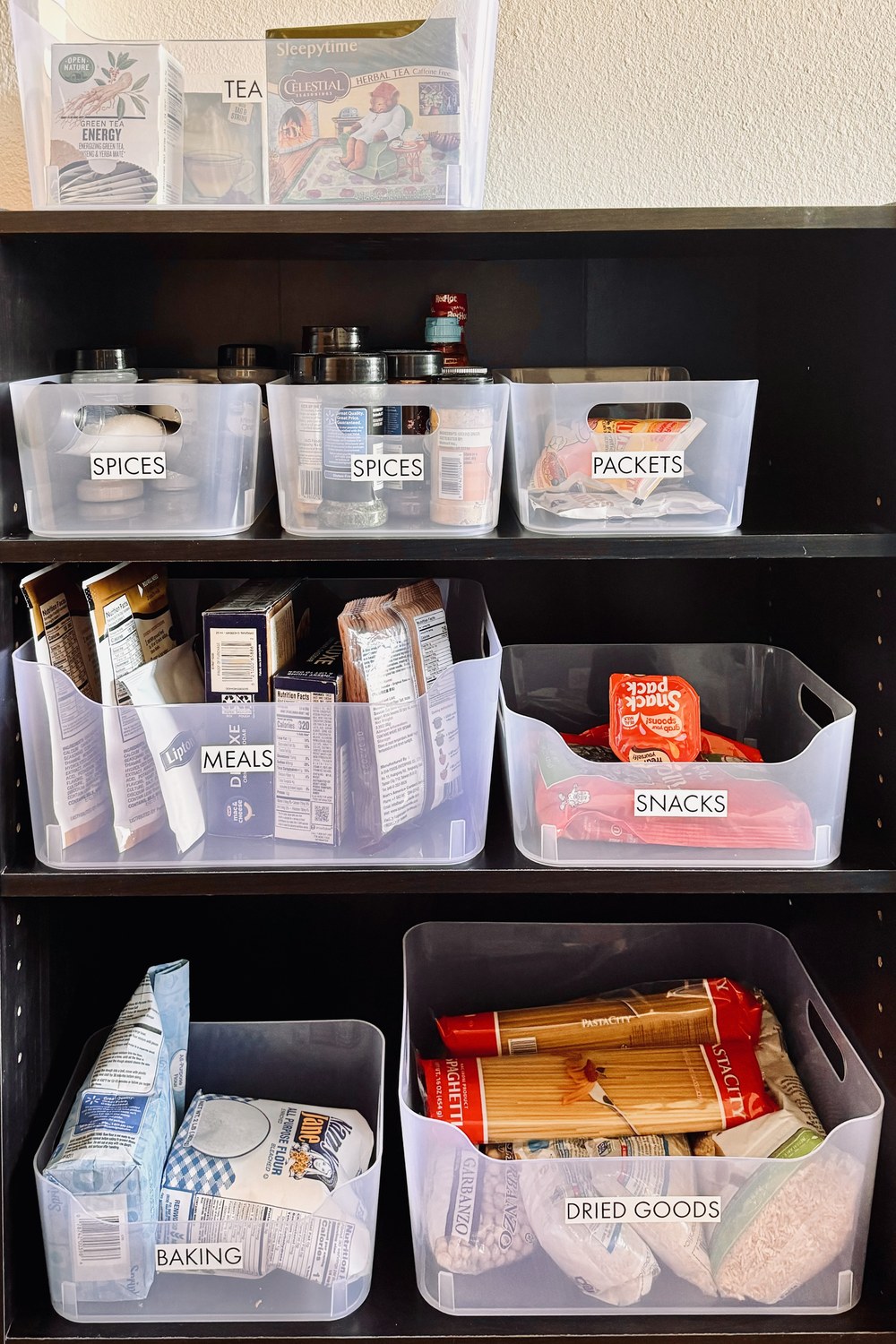 A bookcase pantry with clear bins labeled tea, spices, packets, meals, snacks, baking and dried goods