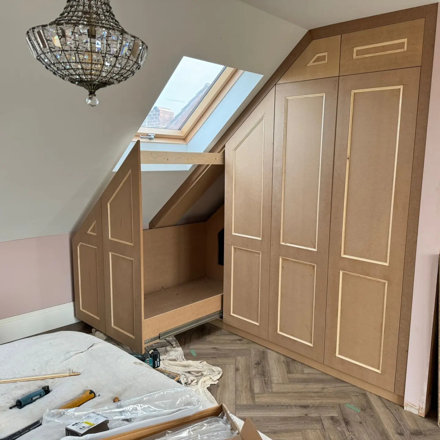 In-progress installation of bespoke fitted wardrobes designed to fit under a sloped ceiling, featuring elegant panel doors and practical interior layout, by a master carpenter in Stourbridge, West Midlands, UK, highlighted by a sophisticated chandelier and a skylight that bathes the room in natural light.