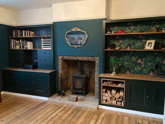 Bespoke dark teal fitted cabinets and shelves by a local West Midlands joiner, featuring an integrated workspace, ornate wallpapered shelving, and a classic wood stove in a rustic fireplace, providing both functionality and elegance to a British home living space with natural wood flooring.